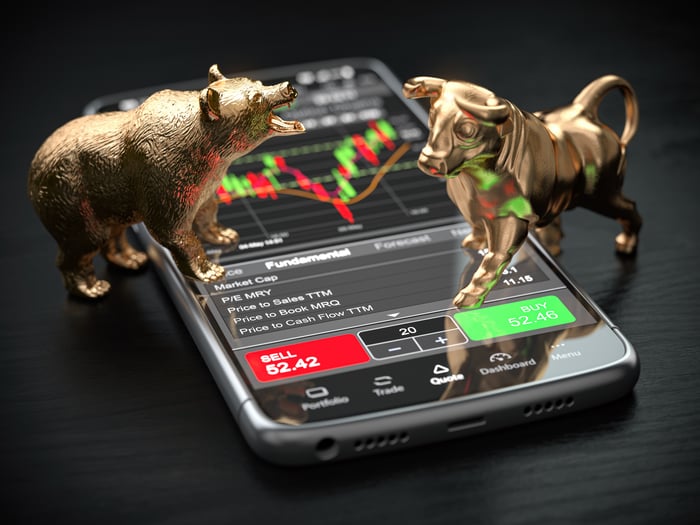 Gold bull and bear figurines placed on top of a smartphone with a stock trading app on the screen.