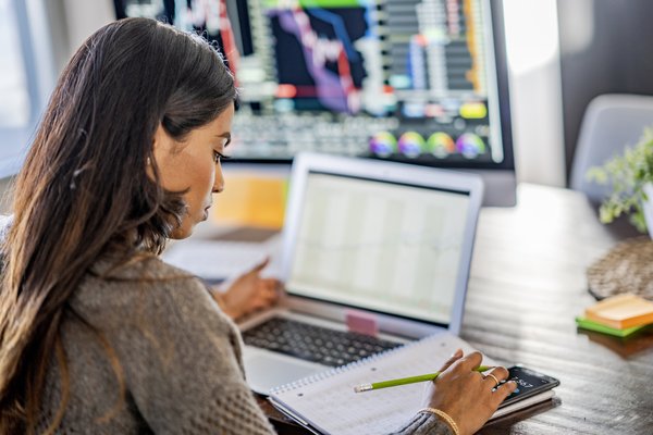 Adult uses calculator in front of laptop next to a larger monitor with charts.