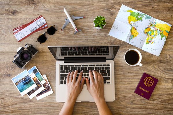 Hands of a person researching travel on a laptop that's sitting on a table with pictures, a passport, brochures, and other travel-related items.